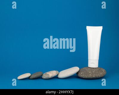 White tube and rounded pebbles on blue background, copy space. Mockup plastic tube for sunscreen or moisturizing cream Stock Photo
