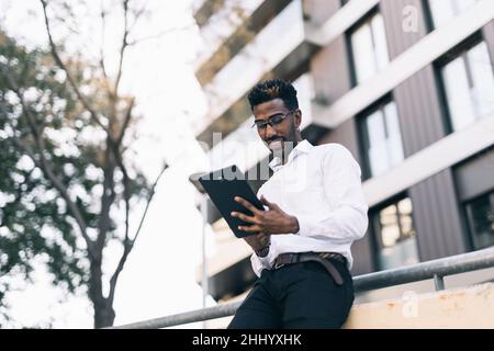 Smiling young black businessman in white shirt working on his tablet in the street Stock Photo