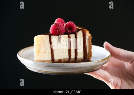 Slice of cheesecake with chocolate sauce on plate in female hand, black background