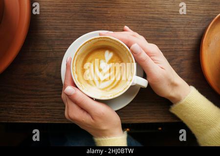 Woman hands in yellow sweater holding cup of fresh coffee with latte art on wooden background. Cappuccino closeup. Coffee break. Hygge concept. Stock Photo