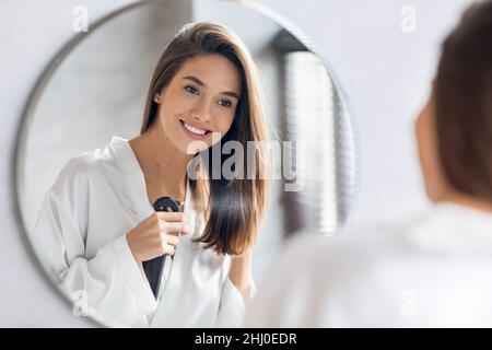 Beautiful Young Woman Applying Hair Spray On Ends Near Mirror In Bathroom Stock Photo