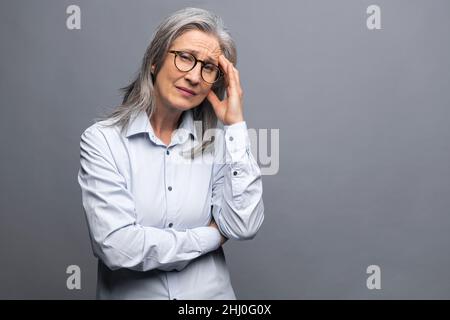 Tired mature female employee feel bored and tired. Upset senior gray-haired woman looking at the camera and feeling headache and burnout isolated over the grey wall Stock Photo