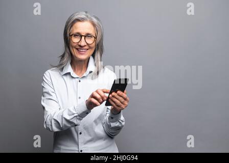 Happy business woman in formal shirt touching to the smartphone screen while feeling great with new phone features. Indoor studio shot isolated on grey background Stock Photo