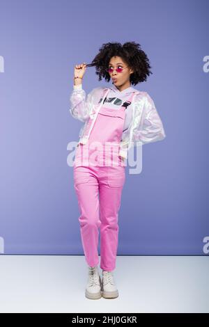 full length of african american woman in pink sunglasses and overalls pulling curly hair and posing with hand on hip on purple Stock Photo