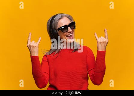 Happy crazy elderly mature woman in sunglasses and headphones making horn signs isolated on yellow. Overjoyed senior gray-haired female in red jumper showing rock symbol, laughing and singing Stock Photo
