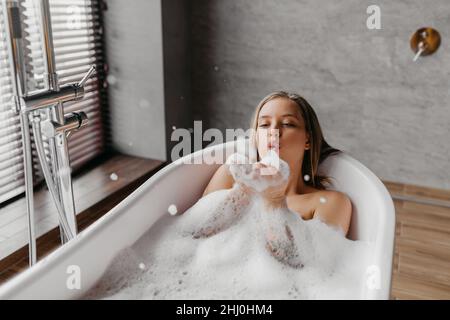 Young woman blowing soap bubbles and having fun while lying in bathtub full of foam at home, copy space Stock Photo