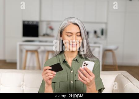 Serene middle-aged grey-haired Asian woman shopping online sitting with credit card and mobile phone at home, multiracial senior 40s female making order online, booking, transfering money Stock Photo