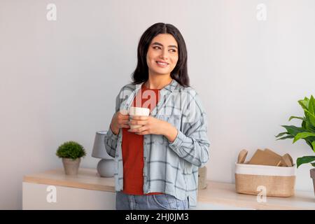 Pretty young Arab woman in casual wear holding cup of fresh coffee, looking aside at home Stock Photo