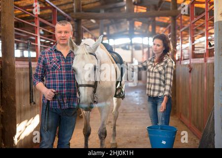 Couple of farmers with bucket before horse standing at stabling Stock Photo