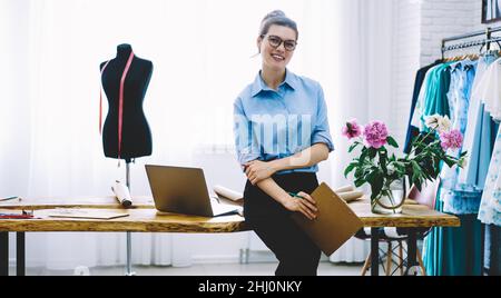 Positive woman with clipboard and pencil in workshop of dressmaker Stock Photo