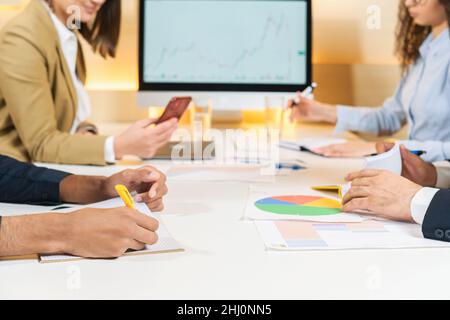 Analyzing important information. Close-up of young professional multiracial managers is sitting at the table with modern laptop while taking notes in the copybook Stock Photo