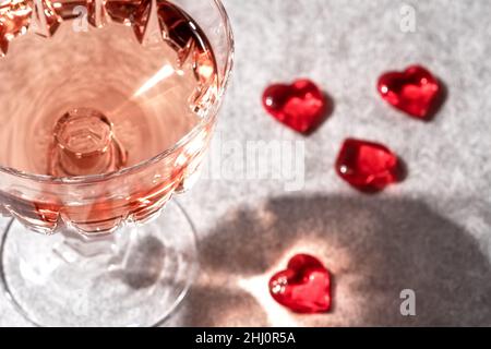 Top view of crystal glass of rose sparkling wine or champagne and red glass hearts on grey stone background in hard light with shadows. Happy Valentines Day concept. Stock Photo