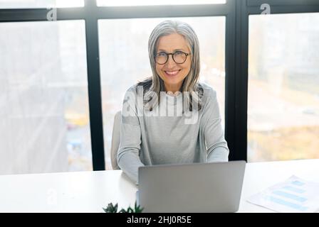 Confident mature female manager ceo sitting at desk indoor, using laptop, gray-haired senior businesswoman looking at the camera sits in bright modern office Stock Photo