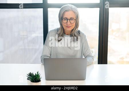 Focused and confident senior gray-haired woman in glasses using a laptop sitting at the desk in office. Concentrated aged woman typing mails answers, messaging online, front view Stock Photo