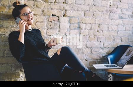 Cheerful young lady speaking on smartphone while drinking hot cappuccino in cafeteria Stock Photo