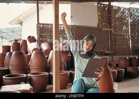 Excited veiled woman raising hand using tableti n pottery shop Stock Photo