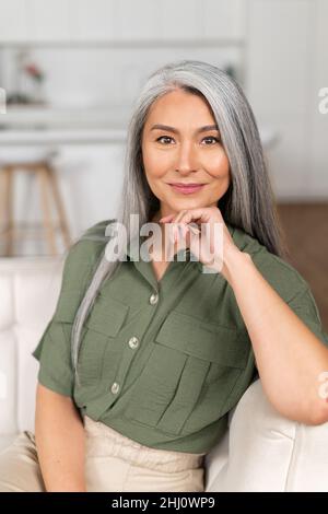 Headshot of smiling good-looking senior Asian female looking at camera. Cheerful carefree grey-haired woman resting at home, vertical shot Stock Photo