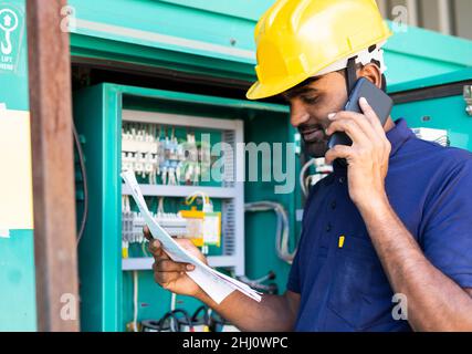 electrical engineer reading or inspecting by talking with colleague on mobile phone in front of circuit board - concept of team work, repair or Stock Photo