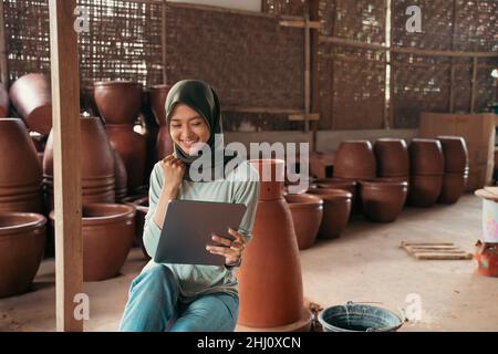 veiled asian woman smiling using tablet while sitting between pottery Stock Photo