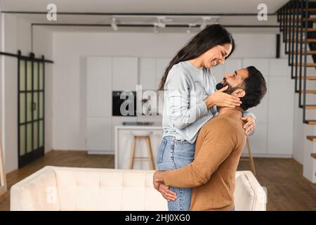 Cheerful Indian guy holding happy woman in arms while standing in apartment. Ethnic latin hispanic couple in love relocating in own flat. Move in new house concept Stock Photo