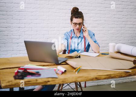 Pensive female tailor talking on smartphone in modern workshop Stock Photo