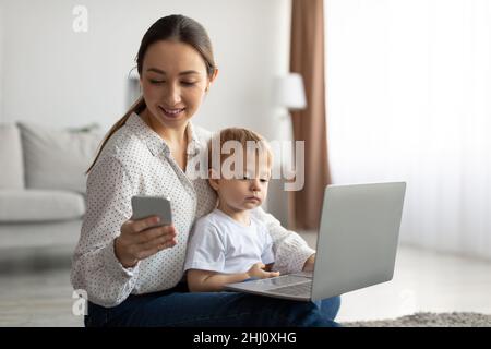 Woman using laptop and smartphone, child boy sitting on mother's knees and watching cartoons online on computer Stock Photo