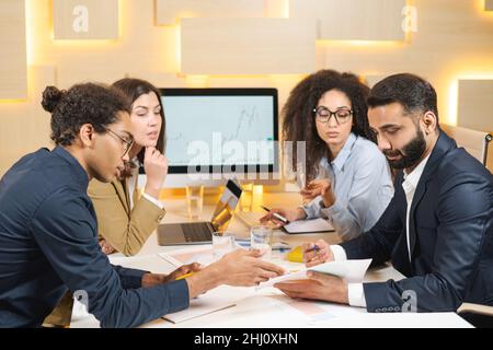 Making great decisions. Young beautiful woman sitting with her colleagues and discussing something with serious expression while her coworkers listening to her sitting at the office table Stock Photo