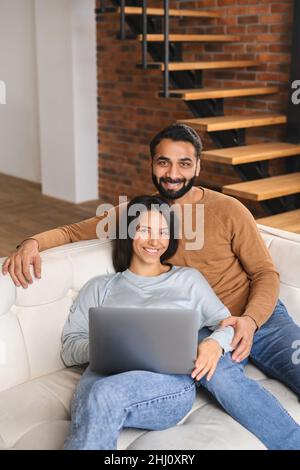 Young Pakistani couple using laptop while sitting on the sofa at home. Spouses watching movie on laptop. Happy man and woman embracing while looking at the camera Stock Photo
