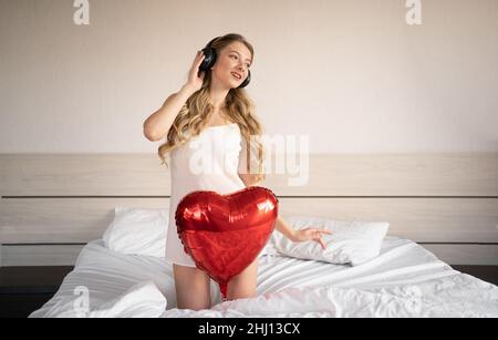Valentine's Day, Women's Day, Anniversary. A young woman in a headphone sits in bed and celebrates February 14 with a red heart-shaped foil balloon Stock Photo