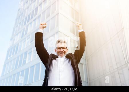 Low angle view of excited senior businessman holding his arms raised and expressing positivity while standing outdoors with office building Stock Photo