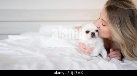 Beauty caucasian woman in pajamas in bed and kissing her cute little brown chihuahua dog Sleeping rest alone on the bed. Banner.