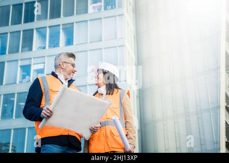 Two engineer colleagues in orange work vests and helmets explore construction documentation. Team concept Stock Photo