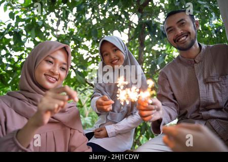 smiling father, mother and daughter lighting fireworks together Stock Photo