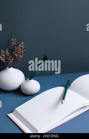 Blank notebook and pencil with vase of dry flowers on desk. navy blue wall background. workspace Stock Photo