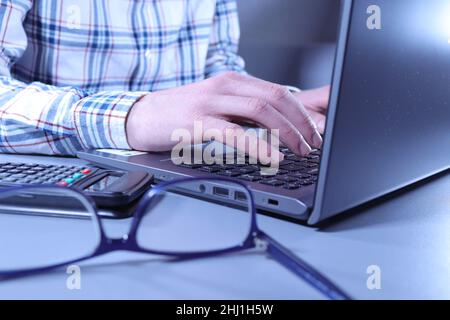 Office worker in checked shirt working at laptop with glasses and calculator on desk Stock Photo