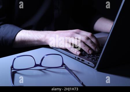 Sinister looking individual in black shirt typing at laptop with glasses on desk Stock Photo