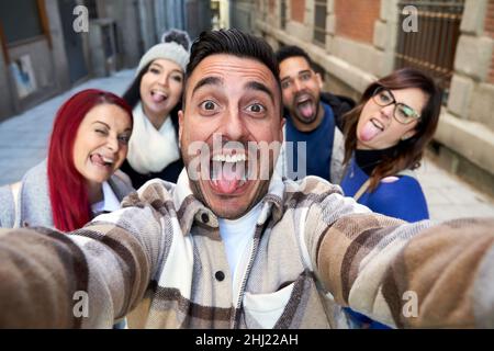Positive multi-ethnic friends standing and making funny gestures while self-portraying on a narrow street and looking at the camera. Selective focus Stock Photo