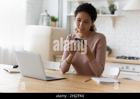 Attractive young 30s Latin woman sit in kitchen with smartphone Stock Photo