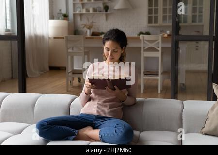 Young woman reading book sit cross-legged on sofa at home Stock Photo