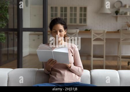 Focused Hispanic woman sitting on cozy sofa reading book Stock Photo