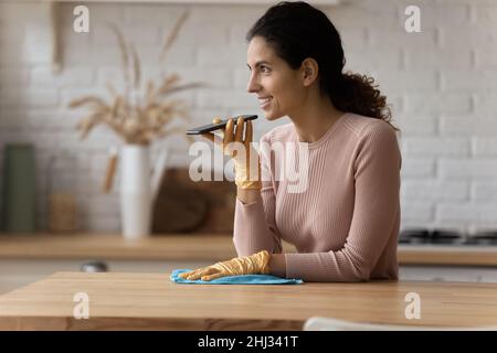 Latin housewife holds smartphone talks on speakerphone while cleaning kitchen Stock Photo