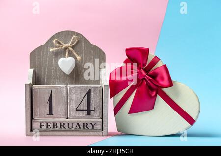 Beautiful heart shaped gift box with ribbon and wooden perpetual calendar on a modern two-tone background. Close-up. Concept of gift for your loved on Stock Photo