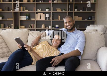 African couple hold tablet relaxing on sofa looking at camera Stock Photo