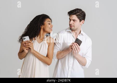 Young multiracial couple frowning while using mobile phones isolated over white background Stock Photo