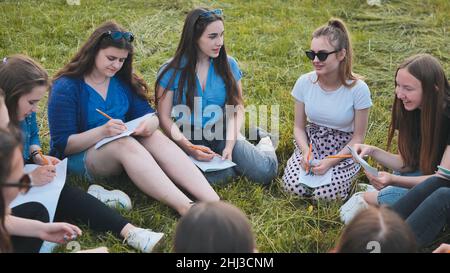 A group of female students are sitting in a circle on a meadow for collective work with notebooks. Stock Photo