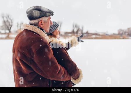 Senior family couple walking outdoors during snowy winter weather. Elderly people enjoying landscape pointing in distance. Valentine's day Stock Photo