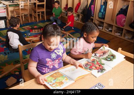 Education Preschool 4 year olds two girls sitting at table and looking quietly at books while group of boys play with train set and tracks in backgrou Stock Photo