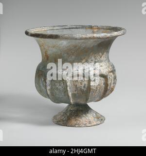 Glass cup with splayed foot 2nd half of 1st century A.D. Roman Translucent blue green.Outsplayed tubular rim, folded over and in; tall curving neck, tapering downwards; thick projecting shoulder; squat, globular body; applied conical foot, formed from a separate gather, with edge cracked-off and ground flat; small, circular, flat bottom.Eleven vertical ribs in high relief tooled out from shoulder down side of body.Rim and body cracked with one hole in side; part of foot broken and missing; few bubbles; dulling, pitting, creamy brown weathering, and faint iridescence.This cup or chalice bears a Stock Photo