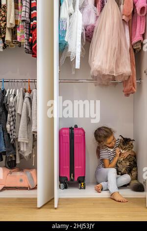 Funny little girl hiding in cupboard hugging cat feeling love and positive emotion Stock Photo