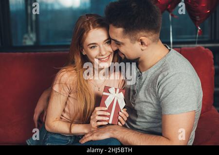 Caucasian couple, girl holding in hands red gift box for anniversary congrats sitting on sofa. Happy birthday. Celebrate party. Valentines day. Helium Stock Photo
